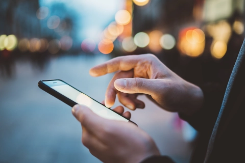 Vor einer Stadtszene sieht der Betrachter eine Hand, die ein Smartphone benutzt. Die tägliche Szene zeigt, wie wenig Angst Nutzer haben, dass das Handy plötzlich anfängt zu brennen.
